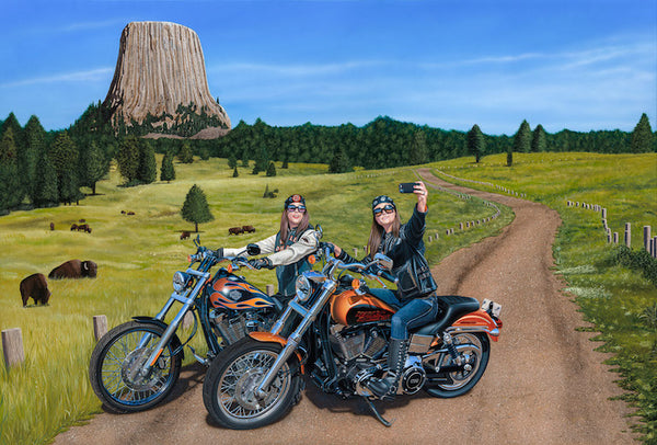 Image of two women writing Harley Davidson motorcycles down a dirt rode, stop to take selfie in front of Devil's Tower in the distance.