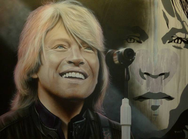 Bon Jovi is pictured with a wide smile on his face, looking in the distance. He is wearing a black leather jacket and a microphone on a white stand is in front of him. In the background is a painted image of his face in various shades of gray.